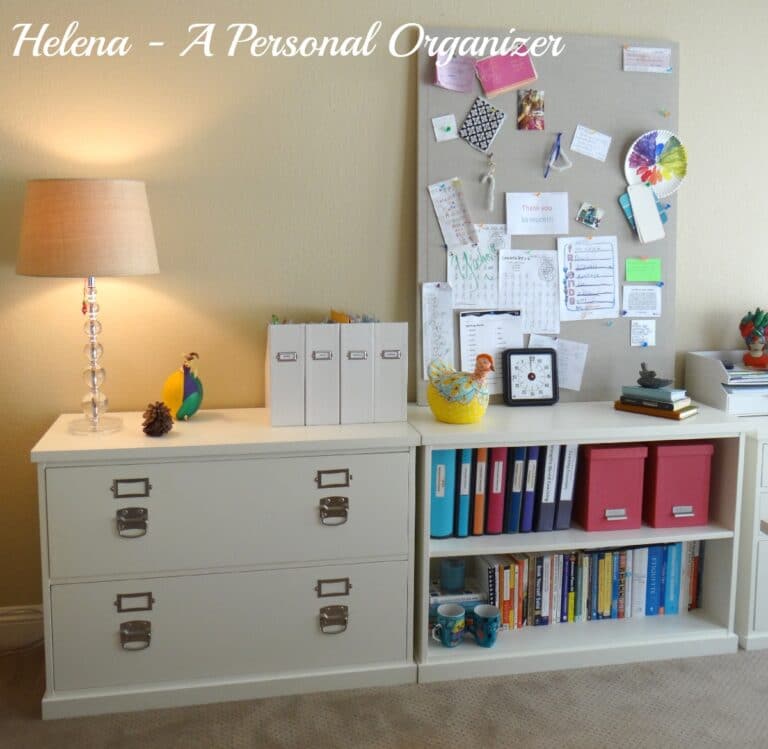         A neatly organized home office nook features a lamp on a filing cabinet, a decorative bird, file boxes, and a corkboard with various notes and decorations. Perfect for back-to-school season, it includes a white shelf with books, binders, and small storage containers to complete the setup.