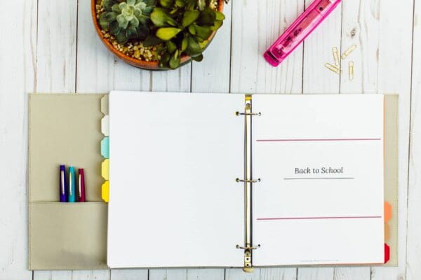 An open beige binder with colorful tab dividers is on a white wooden table. The binder contains a page titled "Back to School". Nearby are a potted succulent plant, a pink ruler, and some paperclips. Pens and markers are in the binder’s pocket, perfect for an organizing printable.