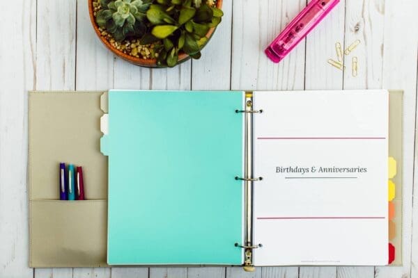 Open binder on a wooden surface, labeled "Birthdays & Anniversaries." Inside the binder are several divider tabs and colorful organizing printables. A potted plant, three pens in varying colors, a pink stapler, and a few paper clips are placed around the binder.