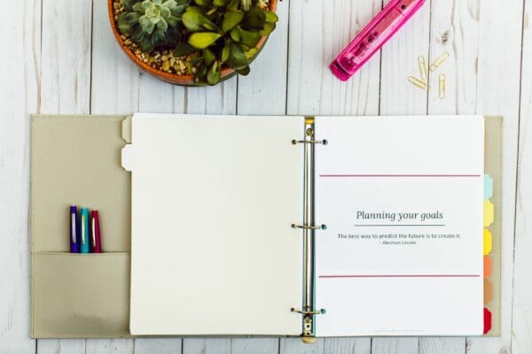 An open planner with labeled tabs reveals a page titled "Planning your goals" with a quote by Abraham Lincoln. Nearby, there are organizing printables, pens, paper clips, a pink ruler, and a pot of succulents on a light wooden surface.