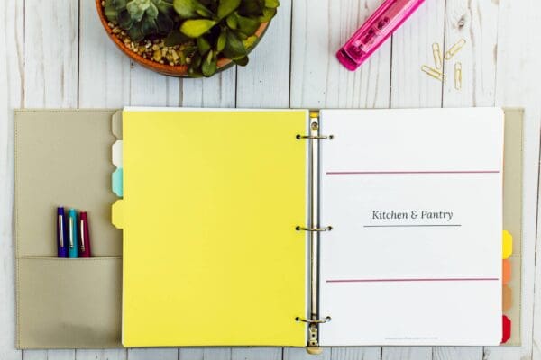 A beige three-ring binder lays open on a white wooden surface. The left side contains colored pens in a pen holder. The right side shows an organizing printable labeled "Kitchen & Pantry." Nearby are a potted plant, a pink stapler, and scattered gold paper clips.