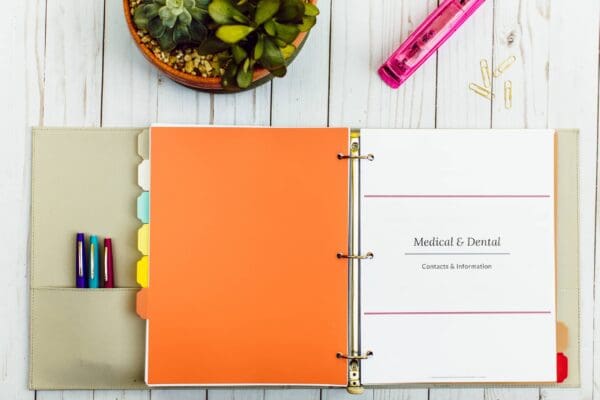 An open binder lies on a wooden surface, showcasing sections labeled with colorful tabs. One visible section is titled "Medical & Dental - Contacts & Information." Nearby is a plant, a pink stapler, and three pens in the binder's pocket. This setup makes organizing printables a breeze.