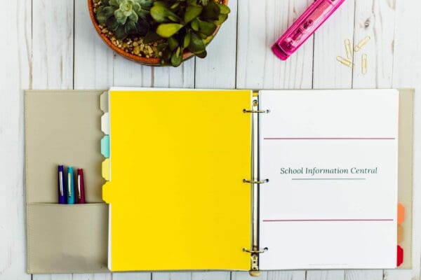 An open three-ring binder with labeled dividers and a cover page titled "School Information Central" sits on a white wooden desk, along with an organizing printable. Nearby are a potted succulent, pink stapler, paper clips, and pens.