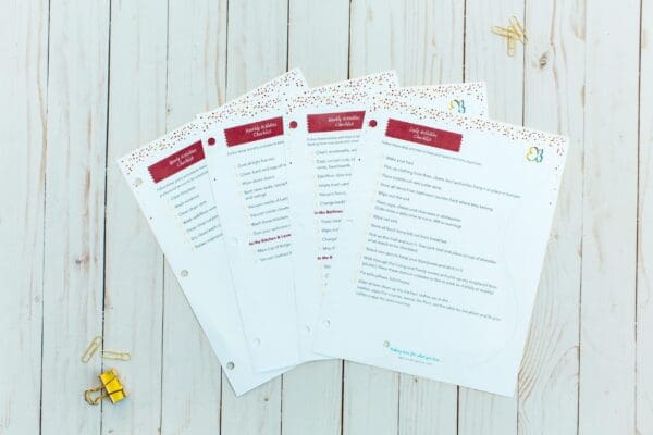 Four organizing printable papers, titled "Holiday Hosting Checklist," placed on a white wooden surface. Each sheet has a decorative header with red and gold accents. Nearby, gold and silver paperclips and a yellow gift box provide a festive feel.