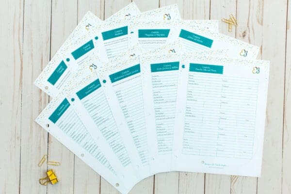 An assortment of organizing printables is laid out on a wooden surface. The sheets, featuring white backgrounds with teal headers, are arranged in a neat overlapping display. Gold paper clips and small decorations add a touch of elegance to the setup.