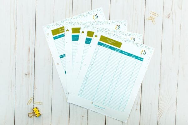 Five sheets of organizing printable financial planner paper are spread out on a white wooden surface. The sheets are labeled "Monthly Expenses" with columns for date, description, amount, and balance. Gold, silver, and small paper clips are scattered around and a gold binder clip is on the left.