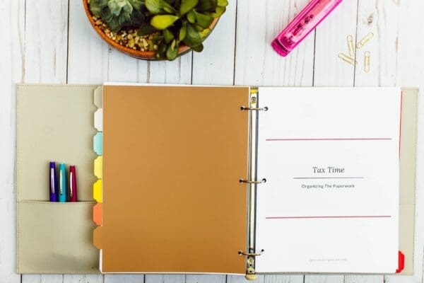 A three-ring binder labeled "Tax Time: Organizing The Paperwork" is open, revealing tabbed sections and a tax checklist. Nearby, a potted plant, pink stapler, and paper clips rest on the white wooden surface. Various pens are tucked into the binder's pocket.