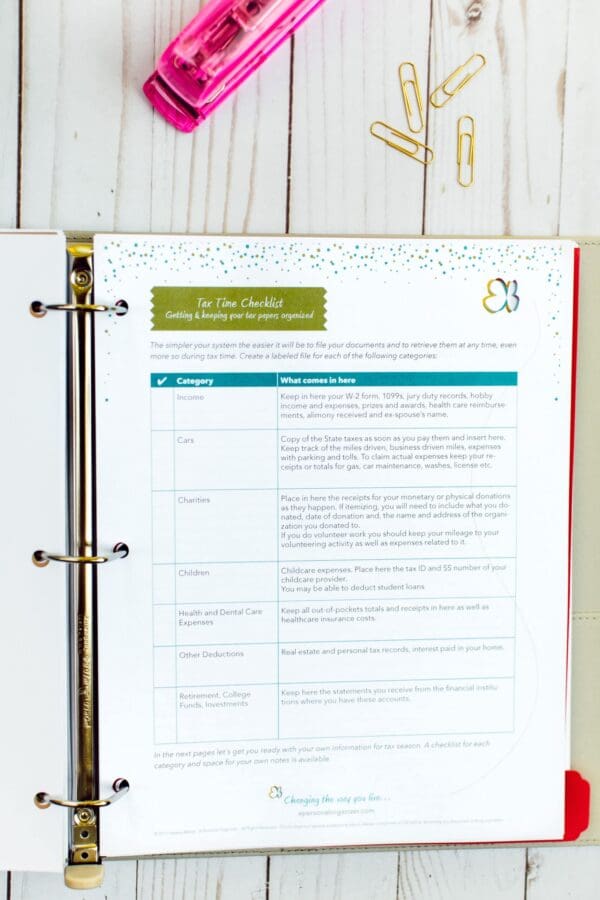 An open binder displays a "Tax Time Checklist" page on a white wooden surface. A pink highlighter and gold paper clips lie next to the binder, which includes categories for income, cars, children, charities, health care, and other deductions—your ultimate tax checklist.