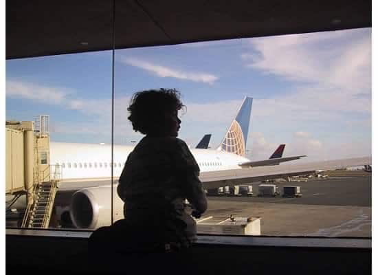 A person with curly hair is silhouetted against a large window at an airport terminal, perhaps imagining the challenges of traveling with kids, as they look out at an airplane parked at the gate. The sky is partly cloudy, and the scene includes parts of the tarmac and airport infrastructure.