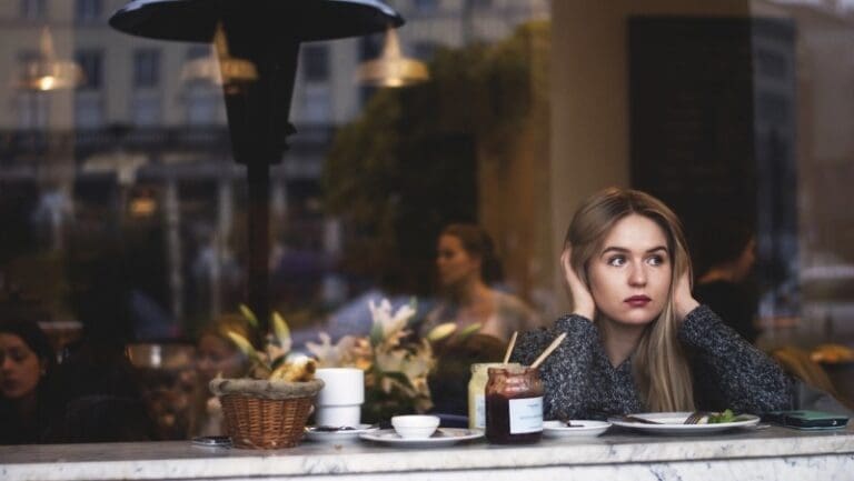 A woman with long blonde hair is sitting alone at a café table, looking contemplative with her hands covering her ears, perhaps pondering how to formalize her thoughts. On the table, there is a basket of bread, a jar of preserves, and some plates. The background shows a busy indoor café environment.