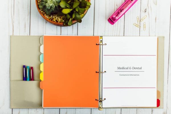 A three-ring binder open on a white wooden surface serves as a comprehensive medical information kit. The binder contains a section titled "Medical & Dental Contacts & Information." Colorful section dividers and various pens fill the side pocket, while a small potted plant, pink stapler, and paper clips sit nearby.