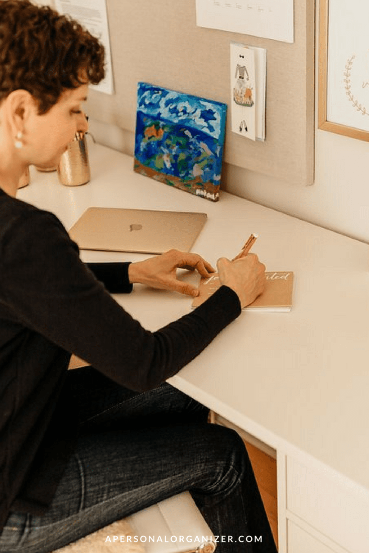 A person sits at a white desk, writing in a notebook. The desk has a closed laptop and a wall with pinned notes and artwork, including a colorful painting and a calendar. The person is focused on their writing, perhaps planning ways to promover como Personal Organizer. The website "apersonalorganizer.com" is at the bottom.