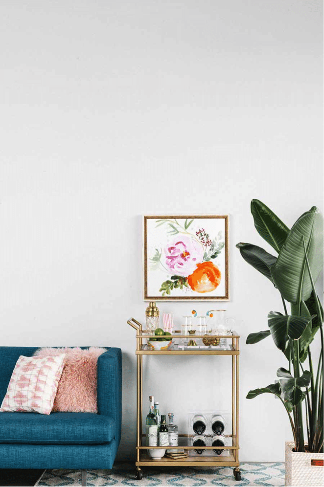 A chic living room corner features a blue sofa with pink cushions, a gold bar cart perfectly styled with drinks and glasses, and a large green plant. A colorful floral painting is hung on the white wall above the bar cart. A patterned rug is partially visible.