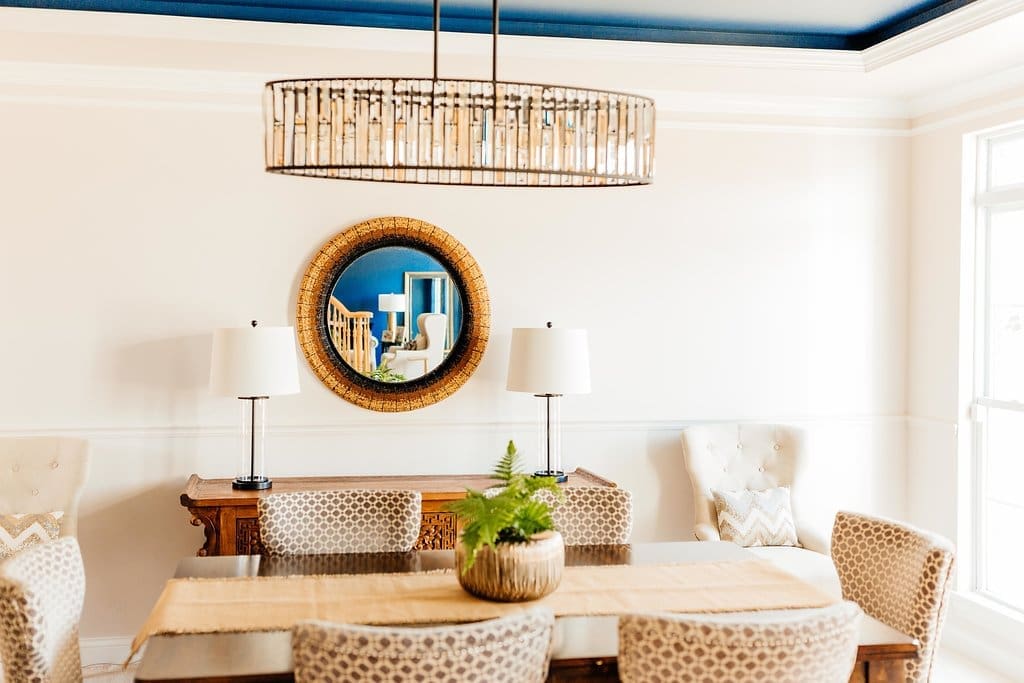 Step into this bright dining room featured in our home tour, showcasing patterned chairs around a wooden table, a plant centerpiece, two white lamps on a sideboard, a round mirror, and a modern chandelier on a blue-trimmed ceiling.
