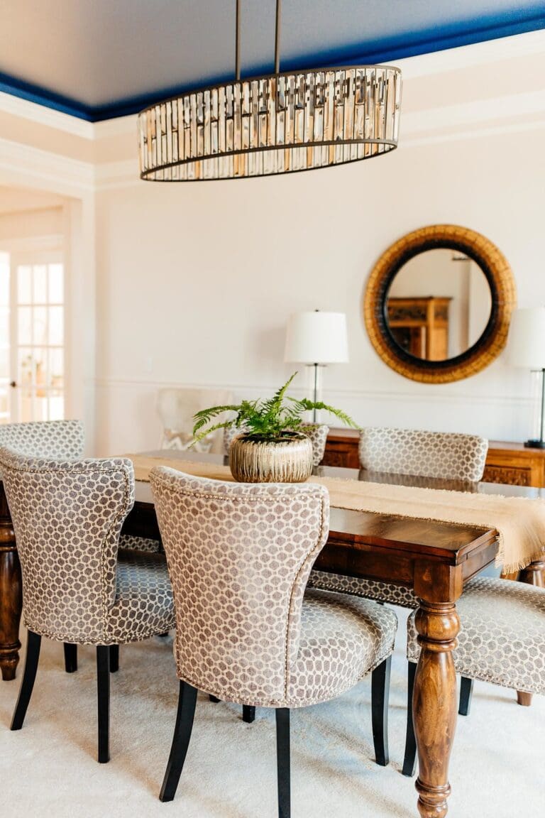 A dining room featured in a recent home tour showcases a wooden table, patterned upholstered chairs, a potted plant centerpiece, a round wall mirror, two lamps, and a modern circular chandelier. Light walls complement the blue accent ceiling.