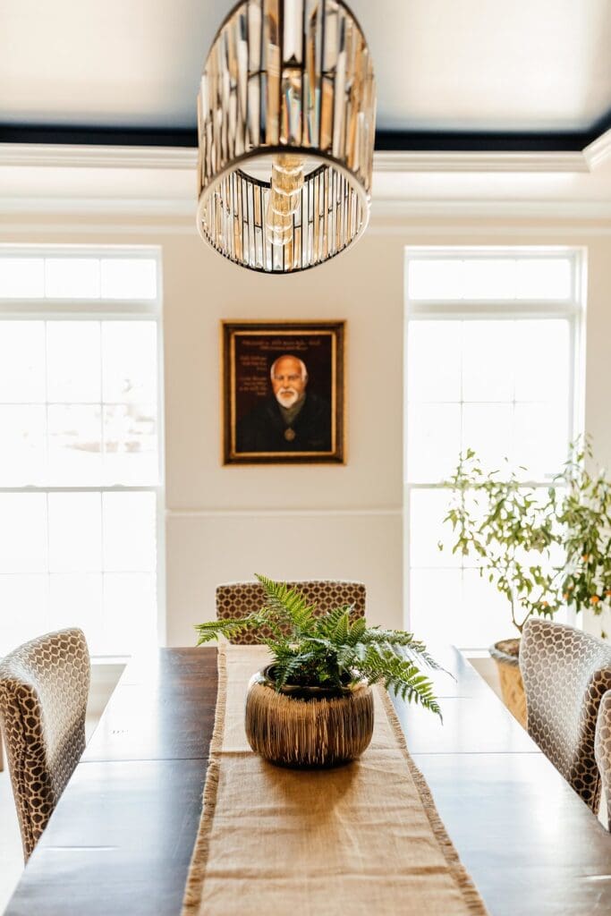A dining room featured on a home tour with a wooden table, patterned chairs, a potted fern centerpiece, large windows streaming natural light, a modern chandelier, and a framed portrait of an elderly man on the back wall.