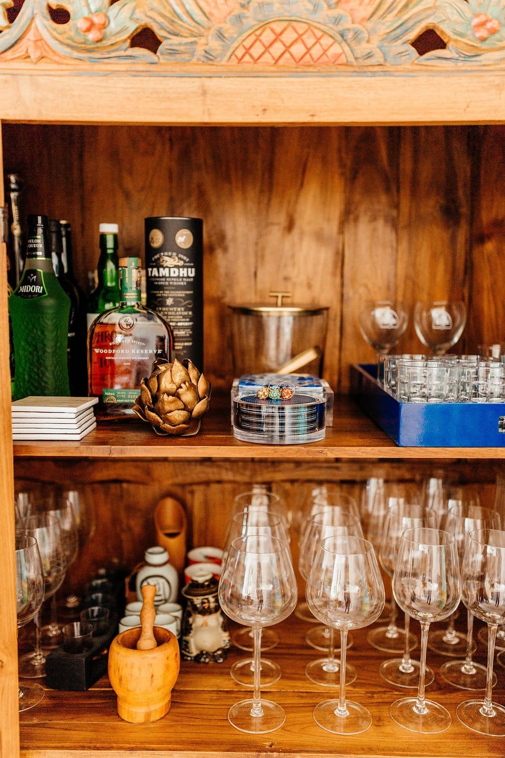 A wooden bar shelf, featured in our latest home tour, displays bottles of liquor, stacked coasters, glassware, a gold artichoke ornament, an ice bucket, a blue tray with small glasses, and several wine glasses on the lower shelf.
