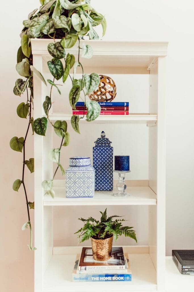A white bookshelf styled for a home tour, featuring plants, patterned blue jars, a gold decorative sphere, a blue candle holder, and stacked books. A leafy plant cascades down from the top shelf.