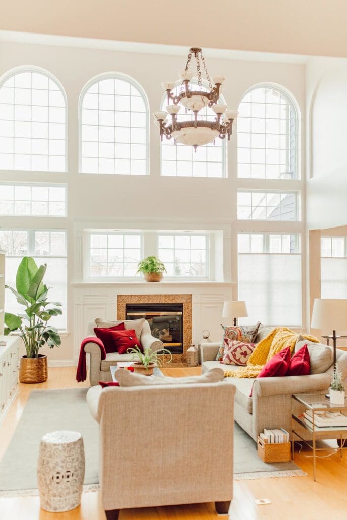 Bright living room with large arched windows, beige sofas with red and yellow pillows, a fireplace, houseplants, and a chandelier. Natural light fills the space, making it perfect for a cozy home tour showcasing colorful decor.