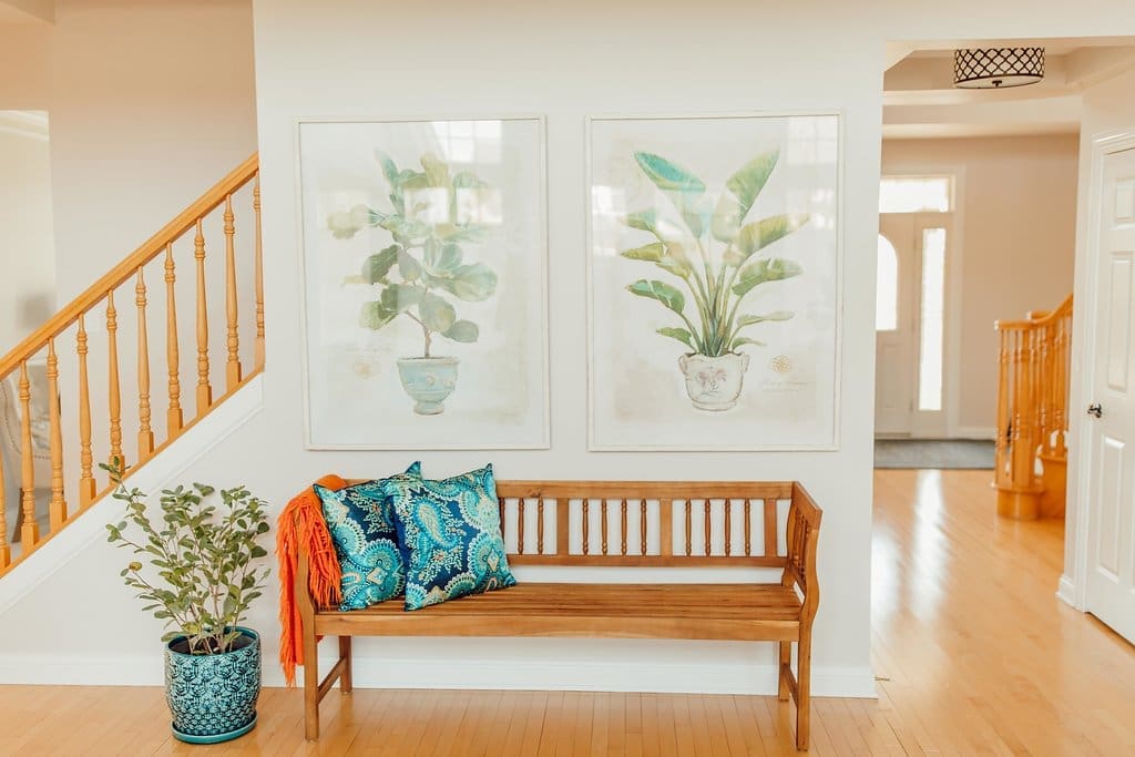 A wooden bench with colorful pillows and an orange throw sits by a staircase in this inviting home tour. A potted plant rests on the floor, while two large botanical prints hang above, all bathed in natural sunlight on light wood floors.