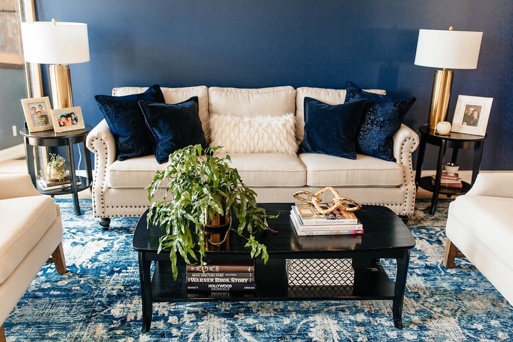 A cozy living room awaits in this home tour, featuring a beige sofa with navy blue pillows and a fluffy white cushion. A dark coffee table holds books, plants, and decor, flanked by white armchairs and gold lamps against a dark blue wall.