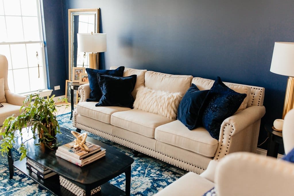 Step inside this home tour—a modern living room with a cream sofa, navy and white pillows, navy blue walls, large window, two lamps, potted plant, blue patterned rug, and sleek black coffee table with books and decor.