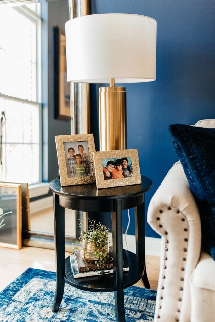 A round black side table holds a gold lamp, two framed family photos, a small potted plant, and books—perfect details for a home tour. The table sits next to a cream upholstered chair in a room with blue walls and a large window.