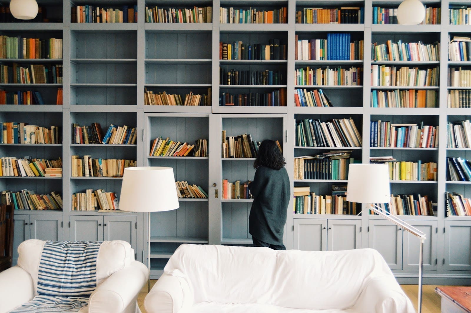 A person stands and looks at books on a large bookshelf filled with various volumes. The bookshelf, an interior design marvel, has multiple compartments and spans the entire wall. A white sofa with a striped throw and two white lamps enhance the cozy ambiance in the foreground.