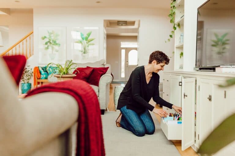 A professional organizer kneels on the floor in a cozy living room, arranging items in a white cabinet. The space features a grey couch with red and green cushions, plants, and sunlight streaming through a door in the background.