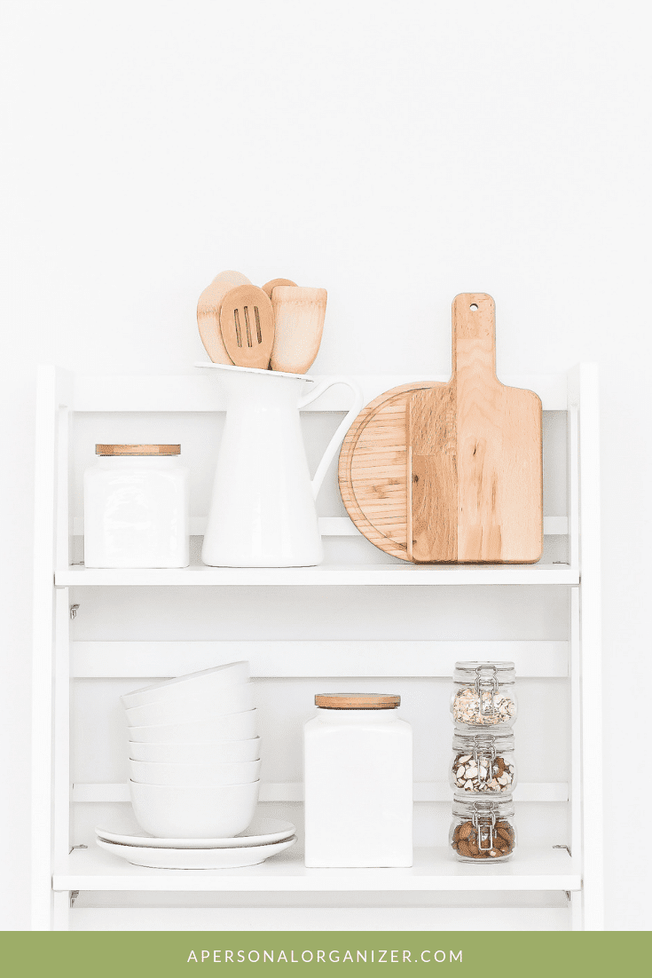 White shelving unit adorned with pantry items, including a ceramic pitcher with wooden utensils, a cutting board, stacked bowls, jars with wooden lids, and a tall jar with dry ingredients. A green banner at the bottom displays "APERSONALORGANIZER.COM.