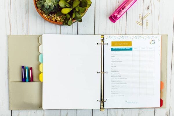 An open beige binder lies on a white wooden surface, displaying a school supplies checklist on the right side. The left side has colorful tabs and three pens in pockets. A pink stapler, gold paperclips, and a pot of succulents are placed nearby, ready to be organized into your cleaning routines.