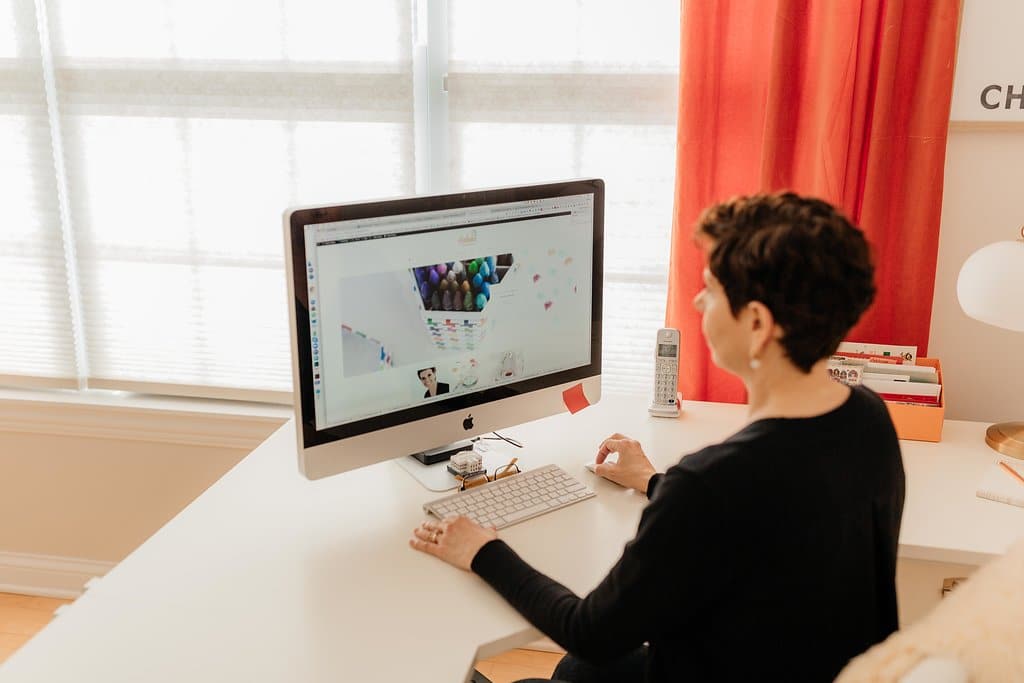 A person with short hair, dressed in a black shirt, sits at a white desk working on an iMac. The desk is well-organized with a keyboard, mouse, cupcake, and landline phone meticulously placed. The room features red curtains, large windows, and files neatly arranged in holders—perfect for efficient office organizing.
