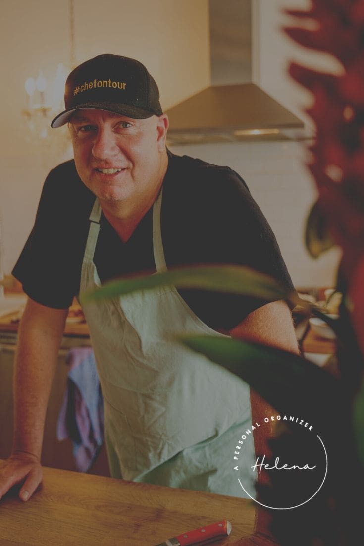 A smiling man in a black "#chefontour" hat and light blue apron stands in a kitchen, perhaps hosting a holiday party. The scene is softly lit with a plant in the foreground. "Helena Personal Organizer" logo appears at the bottom.