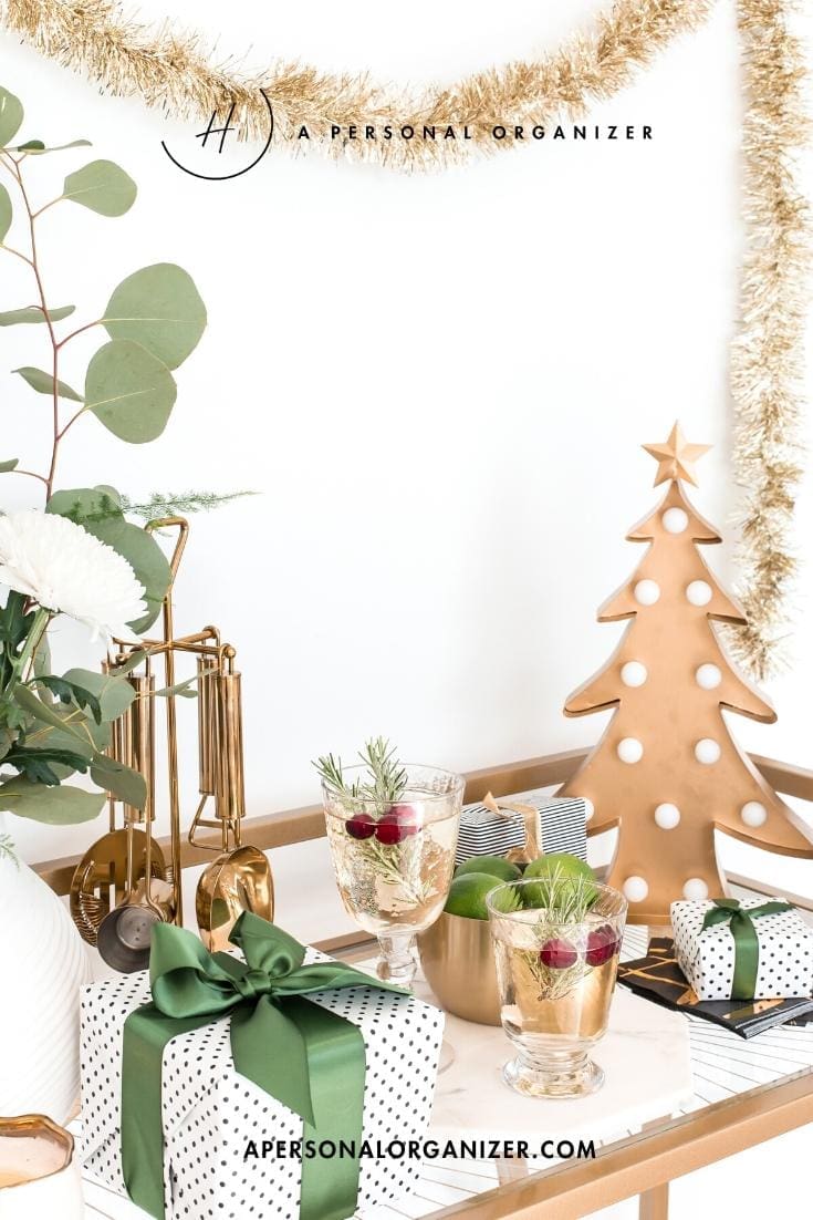 A festive holiday prep scene with a gold drink cart decorated with a metallic Christmas tree, sparkling garland, wrapped gifts with green ribbons, white flowers, cocktails garnished with cranberries and rosemary, and lush greenery.