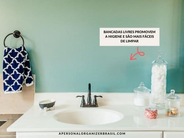 A bathroom counter with a black faucet, a towel featuring a blue and white pattern, and glass jars filled with cotton balls and other essentials sits neatly arranged. A text in Portuguese highlights the advantages of using an organizador de banheiro to keep surfaces clear for easier cleaning.