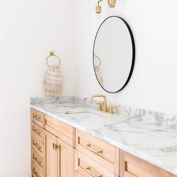 A bathroom with a light wood vanity featuring marble countertops and gold fixtures, complemented by organizing products neatly arranged. A round mirror with a black rim hangs above, and a striped hand towel is on a gold ring holder to the left. The walls and decor remain minimalistic.