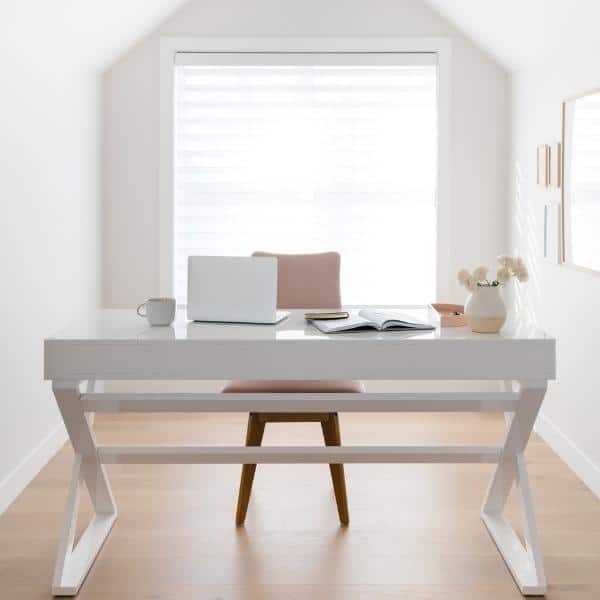 Minimalist home office featuring a white desk and chair, complemented by thoughtfully placed organizing products. A laptop, open notebook, and small vase with flowers grace the surface. The bright window and light wood flooring enhance the airy, clean atmosphere.