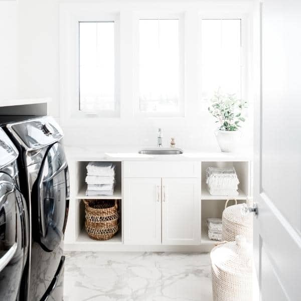 A bright laundry room with white walls and marble flooring features a black washer and dryer. A neatly arranged cabinet holds folded towels, while organizing products like wicker baskets provide stylish storage. A small sink sits nearby, complemented by a potted plant basking in the window light.