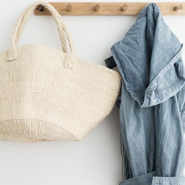 A straw bag and a blue denim shirt hang on a wooden wall rack with multiple pegs, showcasing effective organizing products. The bag on the left and the shirt on the right create a simple, minimalist display.