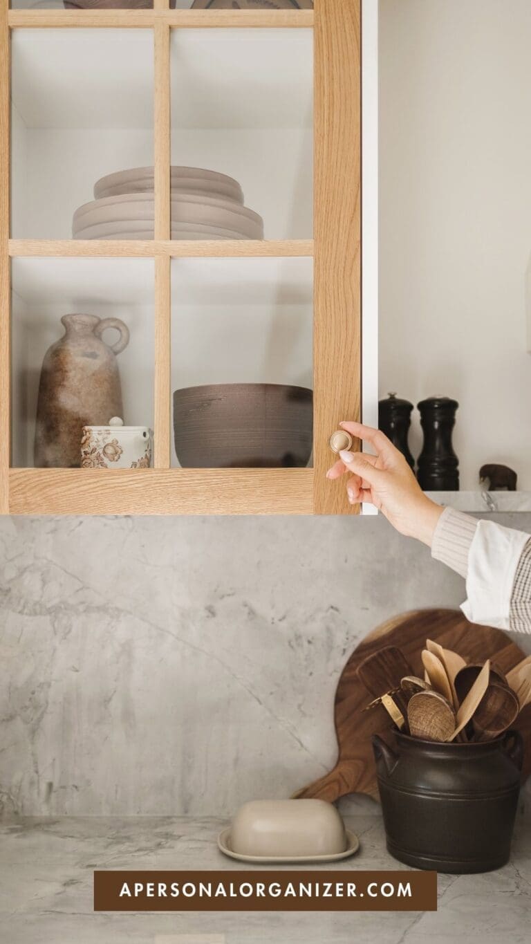 A hand opens a wooden kitchen cabinet with stacked ceramic dishes inside—perfect for decluttering for a move. On the marble counter below are wooden utensils in a container, a butter dish, and cutting boards. Text at the bottom reads "apersonalorganizer.com".