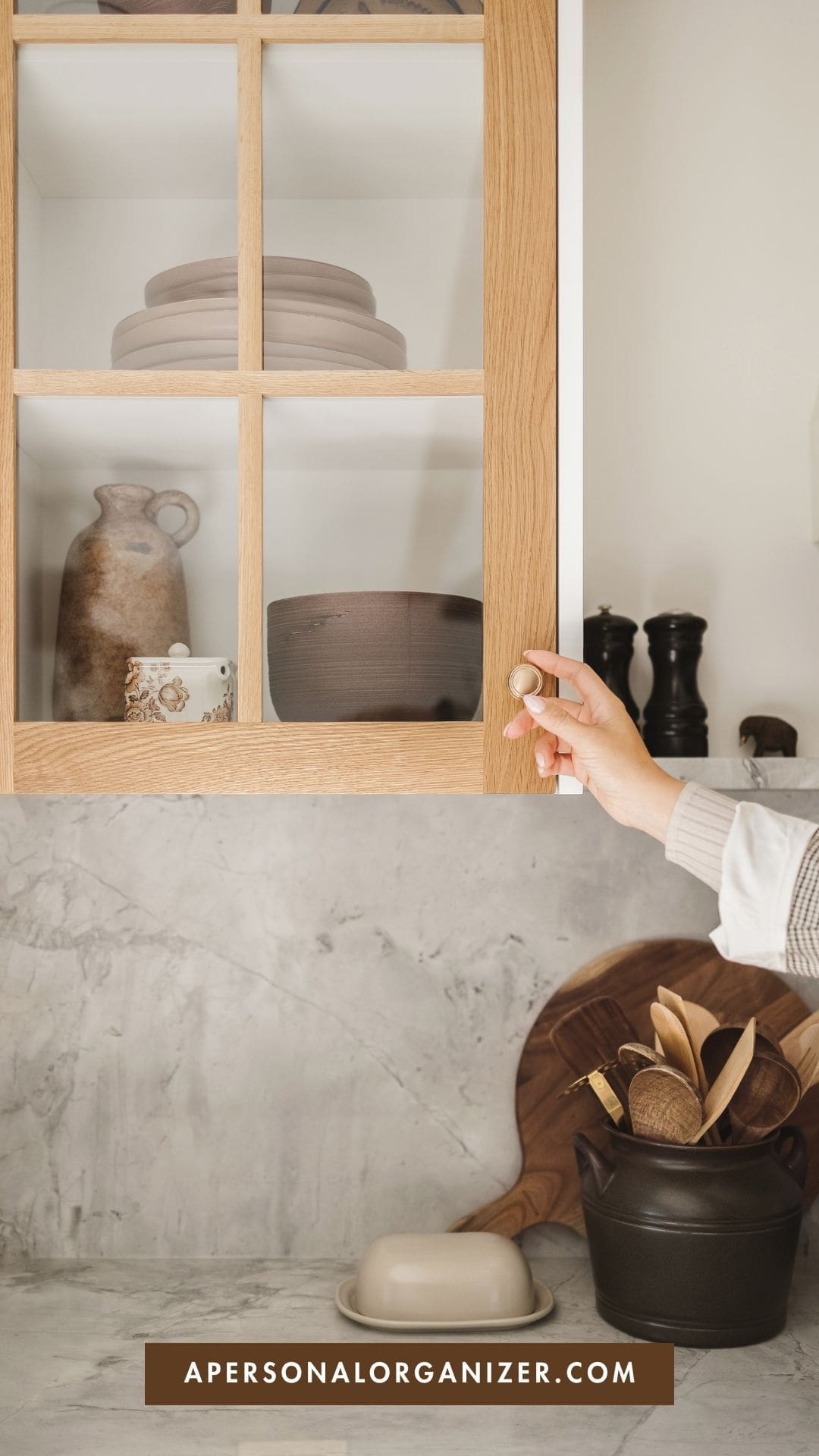 The image shows a person’s hand opening a wooden kitchen cabinet door. Inside the cabinet, there are neatly arranged ceramic dishes and a small pot. On the countertop below the cabinet, there are wooden kitchen utensils in a dark-colored container, along with a butter dish and a wooden cutting board. The text on the image reads: "Decluttering for a Move: A Step-by-Step Guide" with a smaller caption above saying "Home Organization." The website URL "apersonalorganizer.com" is displayed at the bottom of the image. The overall tone is clean and organized, reflecting the theme of home organization.