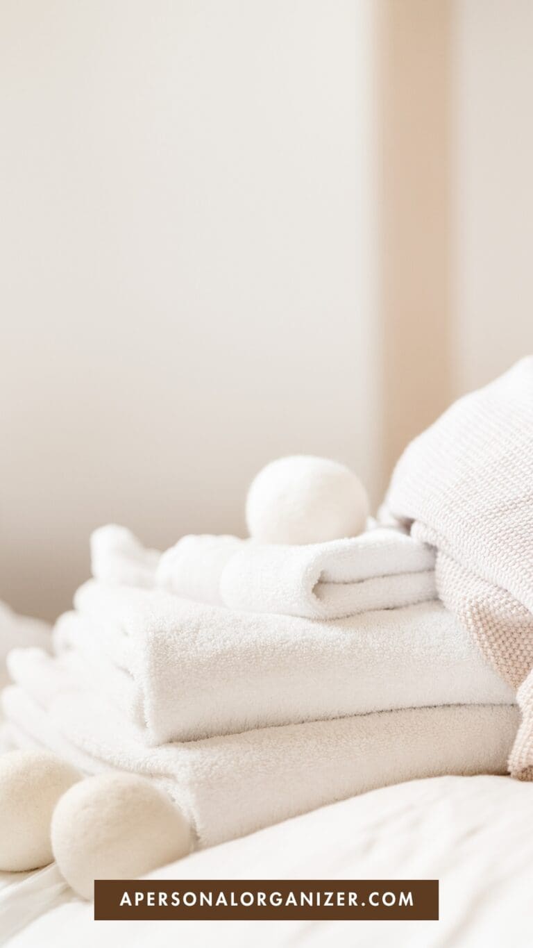 A stack of neatly folded white towels and cozy textiles on a bed, with wool dryer balls on top; a soft, neutral background. The text "APERSONALORGANIZER.COM" appears at the bottom.