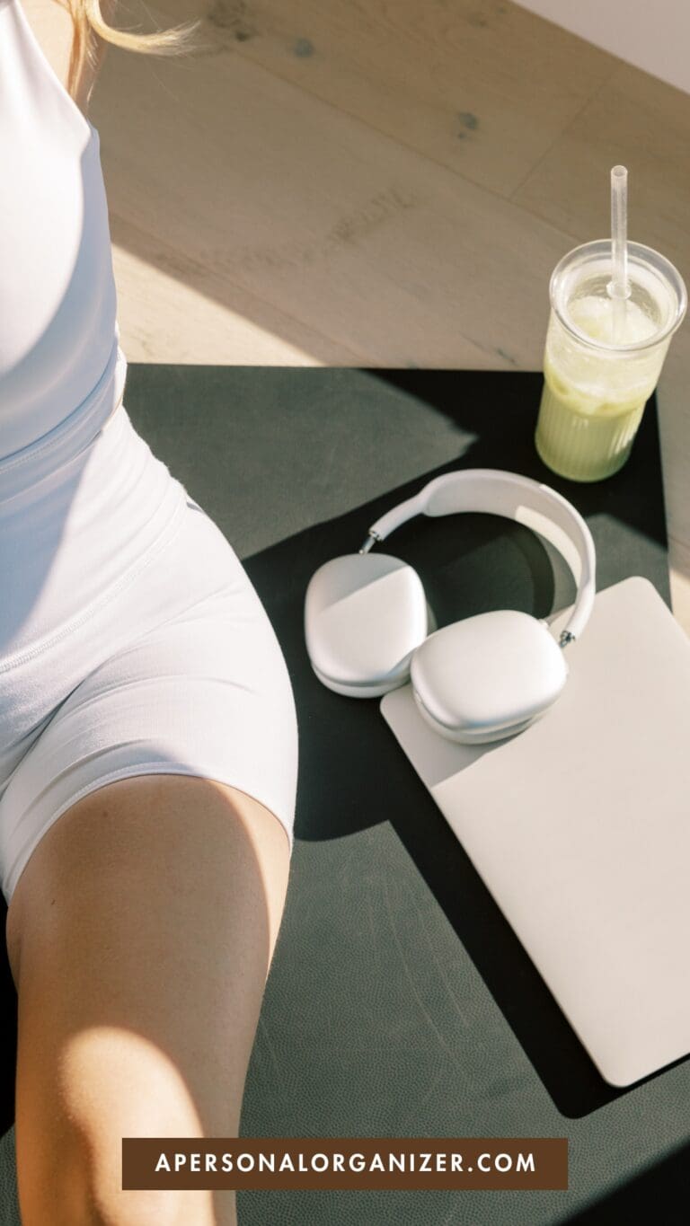 A woman in white workout clothes sits on a black mat next to wireless headphones, a closed laptop, and a green smoothie with a straw. Sunlight casts shadows across the scene. Text reads "APERSONALORGANIZER.COM".