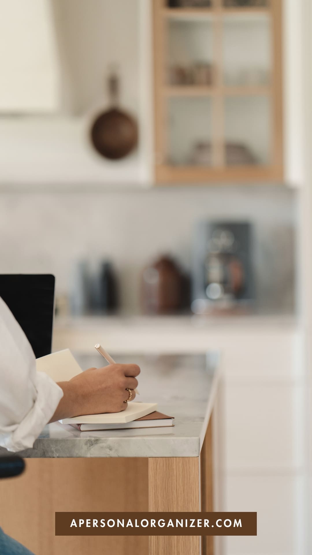 A person sits at a kitchen counter, writing a back to school to do list in a notebook with a pencil. The background shows blurred kitchen shelves and appliances. A label at the bottom reads "APERSONALORGANIZER.COM.