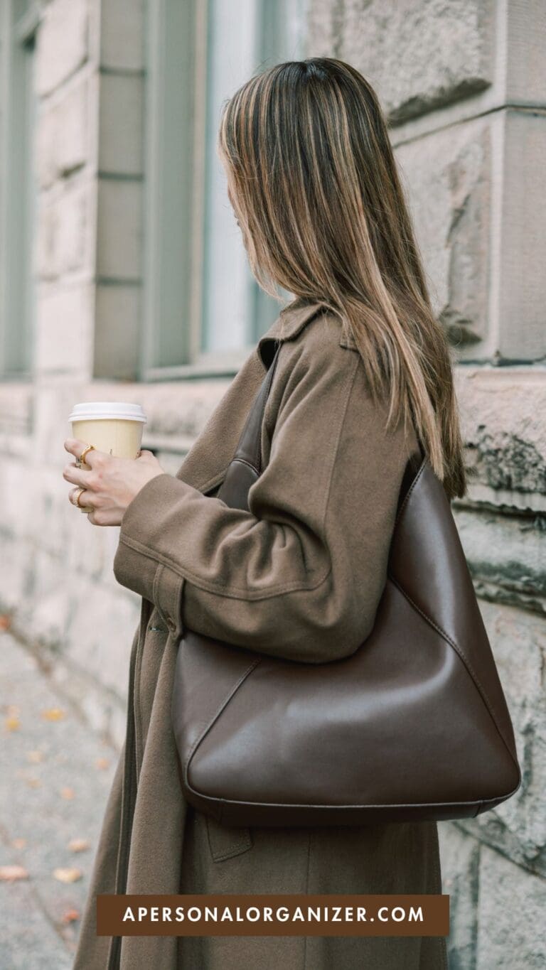 A woman with long hair wearing a brown coat holds a coffee cup and carries a large brown purse—showcasing classic Fall Outfit Ideas. She stands outside by a stone building. The text "APERSONALORGANIZER.COM" appears at the bottom.