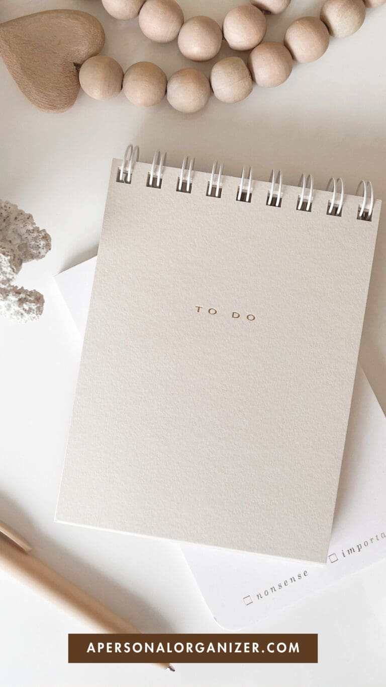 A beige spiral notebook with "TO DO" written on the cover lies on a white surface, surrounded by a pen, a wooden bead bracelet, a heart-shaped object, and a card. Text at the bottom reads "APERSONALORGANIZER.COM".